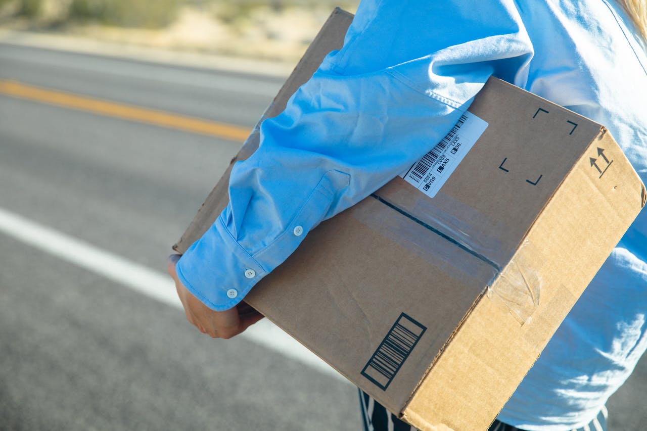 about-us Close-up of a person carrying a cardboard box on a sunny road with a blue shirt.