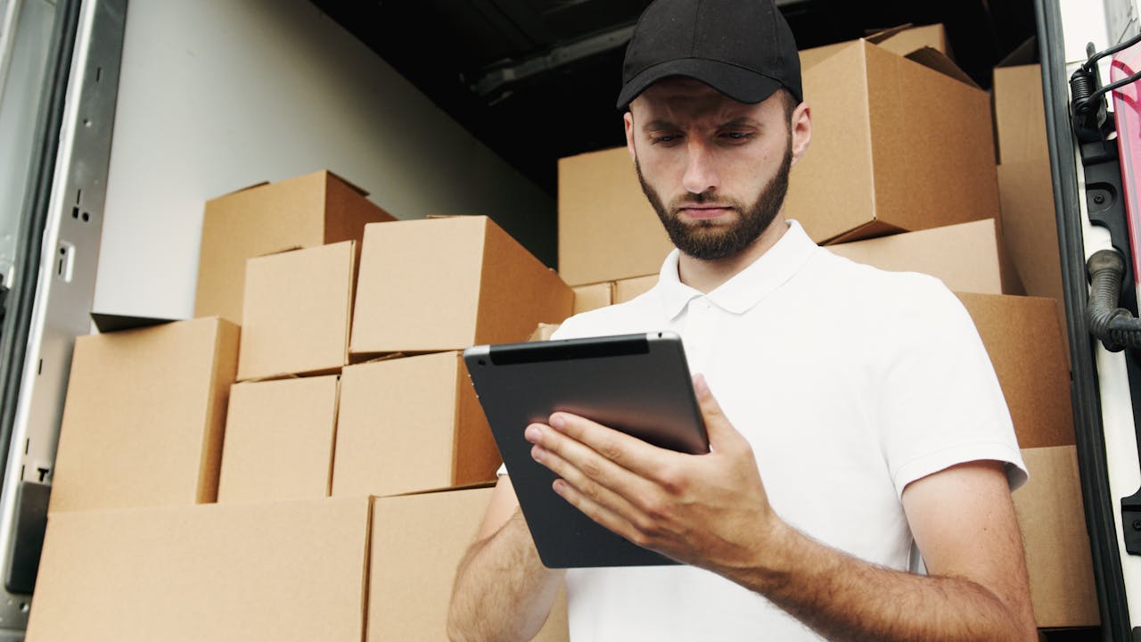 who-we-are Delivery worker using a tablet to manage shipments with stacked boxes in the background.